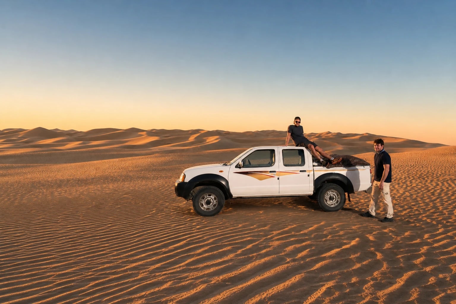 Zwei Teammitglieder mit einem weißen Pick-up auf Sanddünen bei Sonnenuntergang